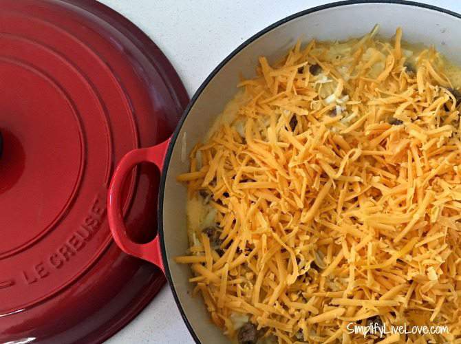 shredded cheddar cheese on top of sausage and hash browns in red baking dish with lid